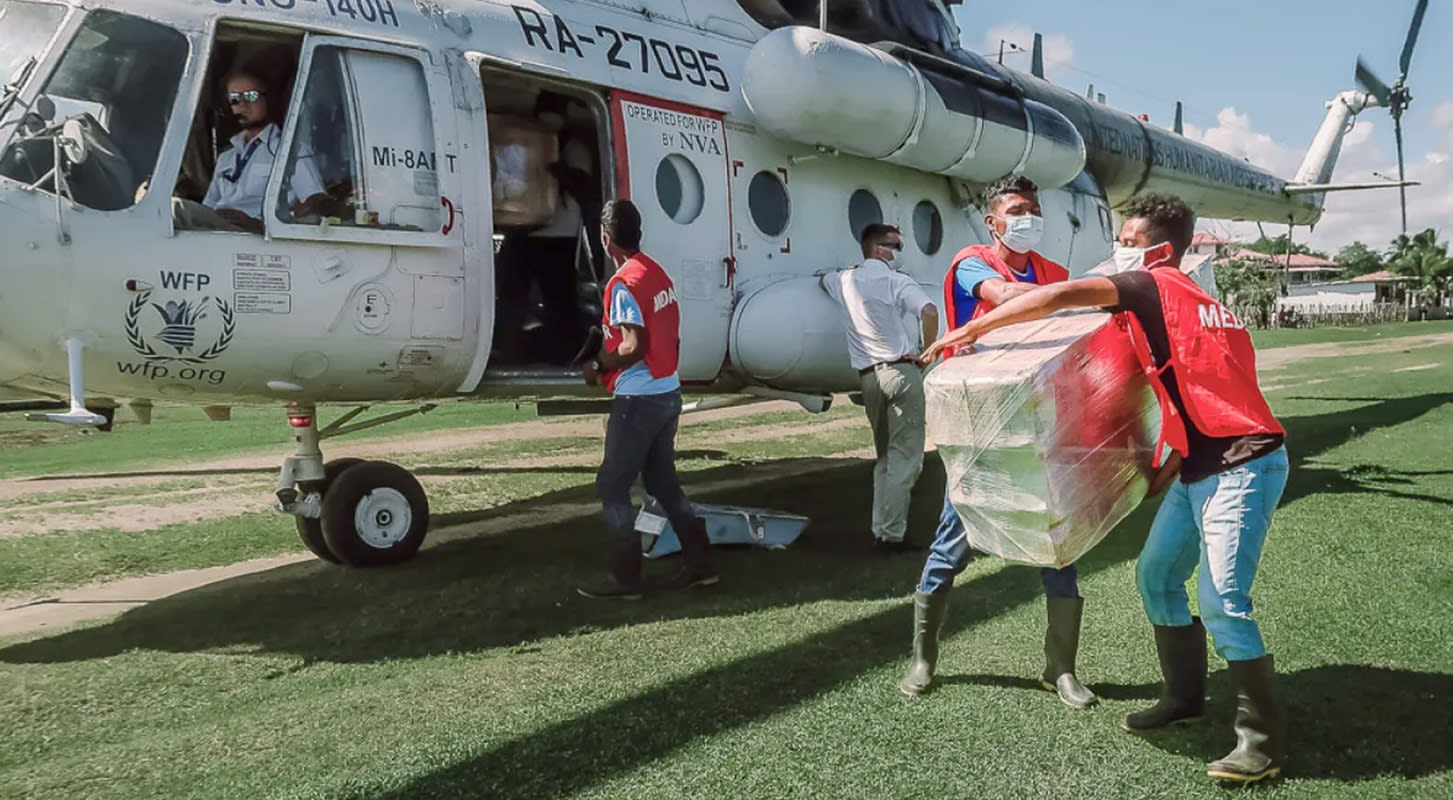 Individuals in red Medair vests load boxes onto a helicopter under a clear sky, preparing for transport to a remote location.